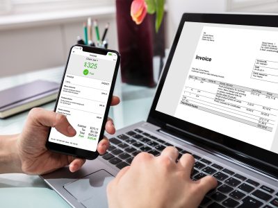 Close-up Of Businessperson Using Laptop While Paying Invoice On Mobilephone In Office